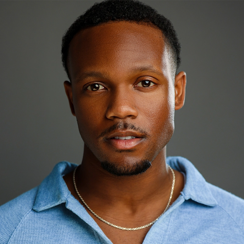 A young man with short, neatly trimmed hair and a goatee poses against a plain gray background. He is wearing a light blue collared shirt slightly open at the neck and a thin silver chain necklace. His expression is calm and confident.