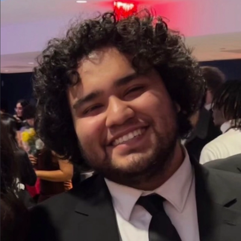 A young man with curly dark hair and a beard smiles broadly. He is wearing a black suit, white shirt, and black tie. The background is softly blurred, showing people in formal wear and warm, ambient lighting.