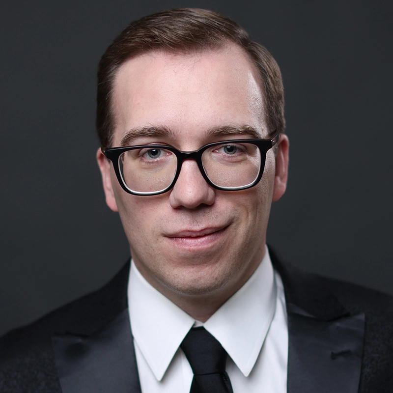 A man with short, light brown hair wears black rectangular glasses, a white dress shirt, black tie, and a black suit jacket. He is smiling slightly and posed against a plain, dark gray background.