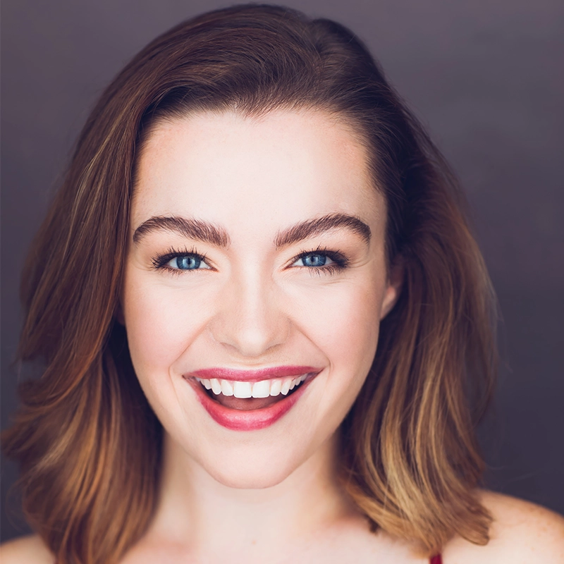 Grace Whetstone, a young woman with fair skin and shoulder-length brown hair, smiles brightly. She has blue eyes, well-groomed eyebrows, and wears natural makeup with pink lipstick. The dark gray background highlights her cheerful expression.