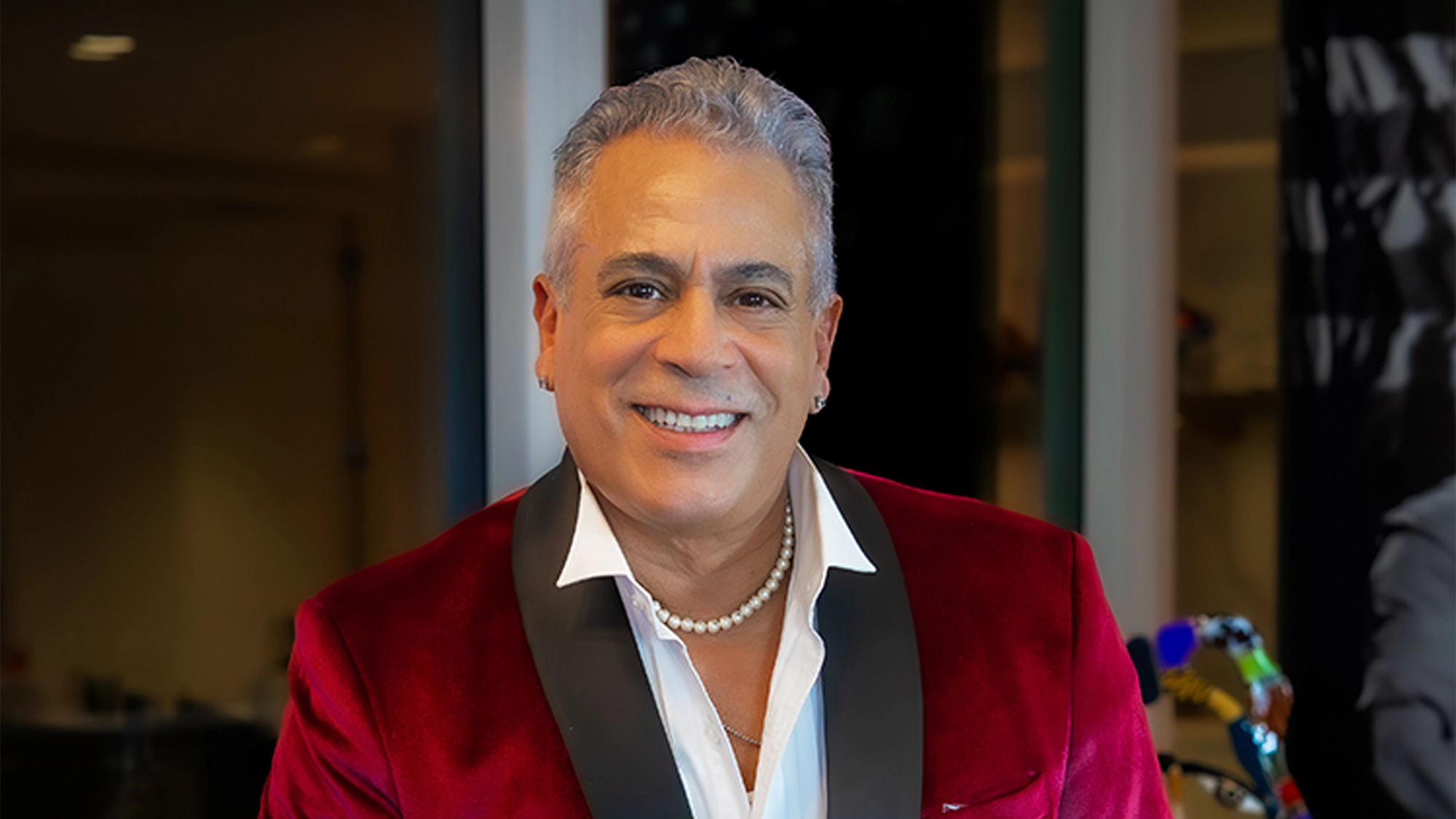 A smiling man with short gray hair wears a red velvet jacket with black lapels, a white dress shirt open at the collar, a pearl necklace, and earrings—capturing the vibrant energy of Cabaret Disco live indoors. Blurred background elements are behind him.