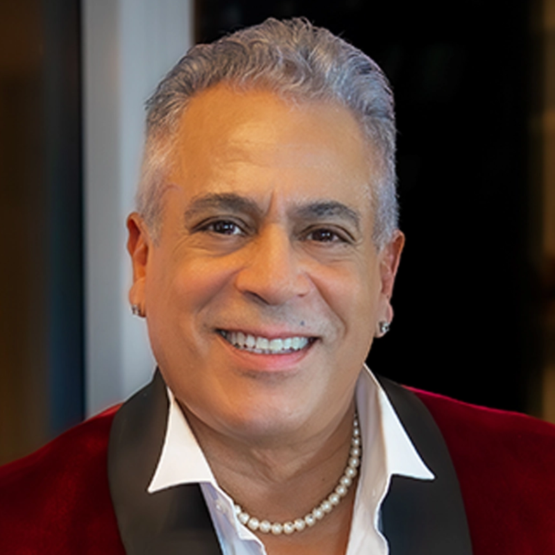 A smiling middle-aged man with short, gray hair wears a red velvet jacket, white shirt, pearl necklace, and hoop earrings. He is looking directly at the camera with a friendly expression, set against a blurred background.