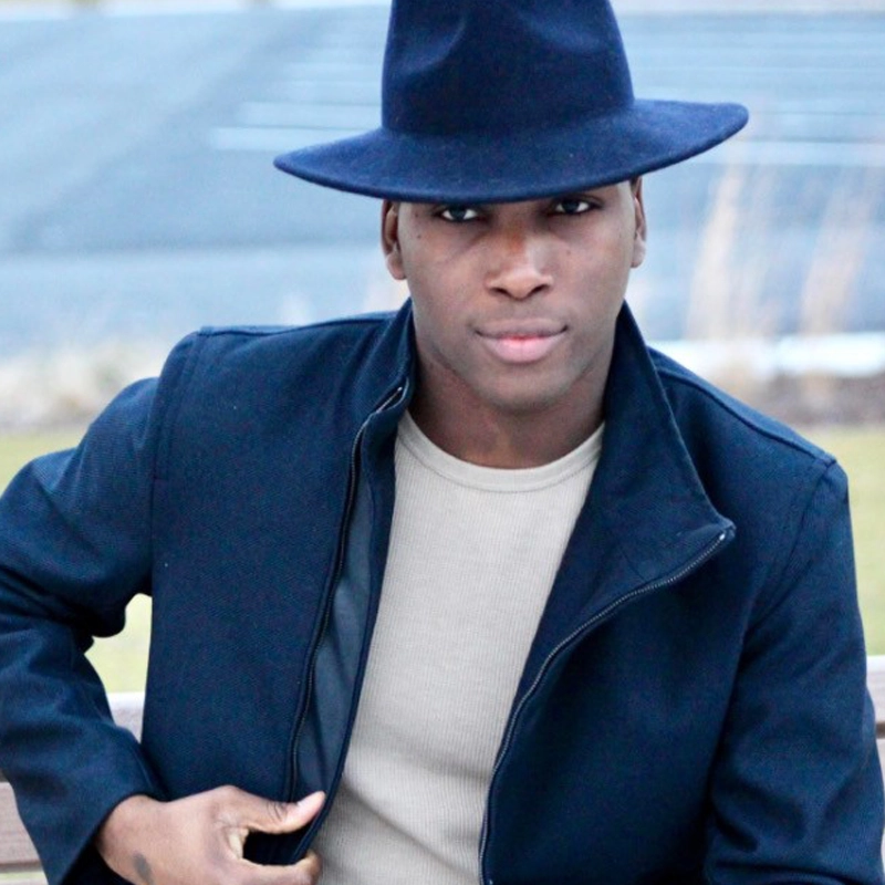 A young man, reminiscent of Travis Finlay, wears a navy blue wide-brimmed fedora, a matching dark jacket, and a light beige shirt as he sits outdoors on a bench, looking confidently at the camera against blurred blue and green hues.