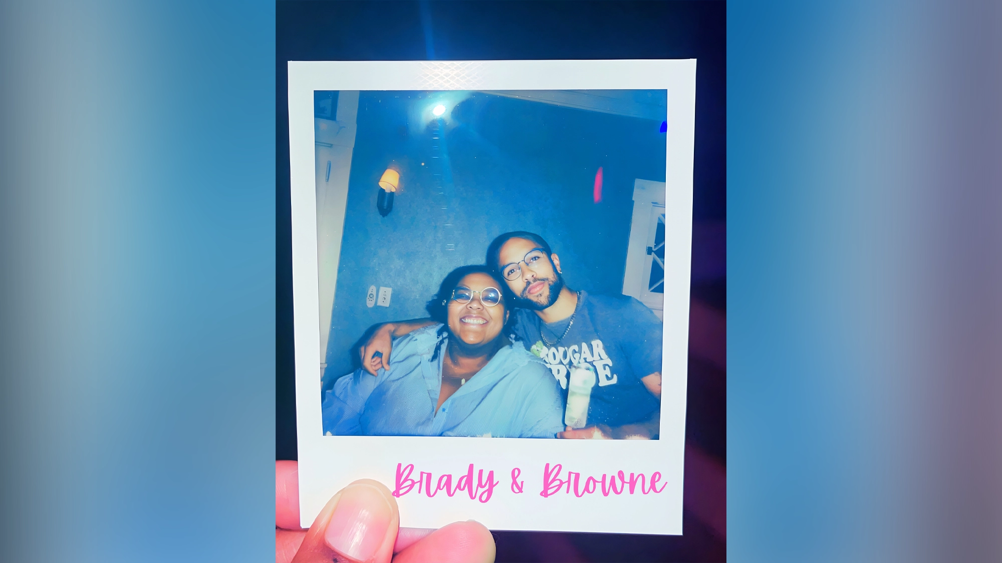 Flame - Site Image A hand holds a Polaroid photo of Xander Browne and Amara Janae Brady smiling indoors. One wears glasses and a blue shirt; the other, in gray, holds a drink.