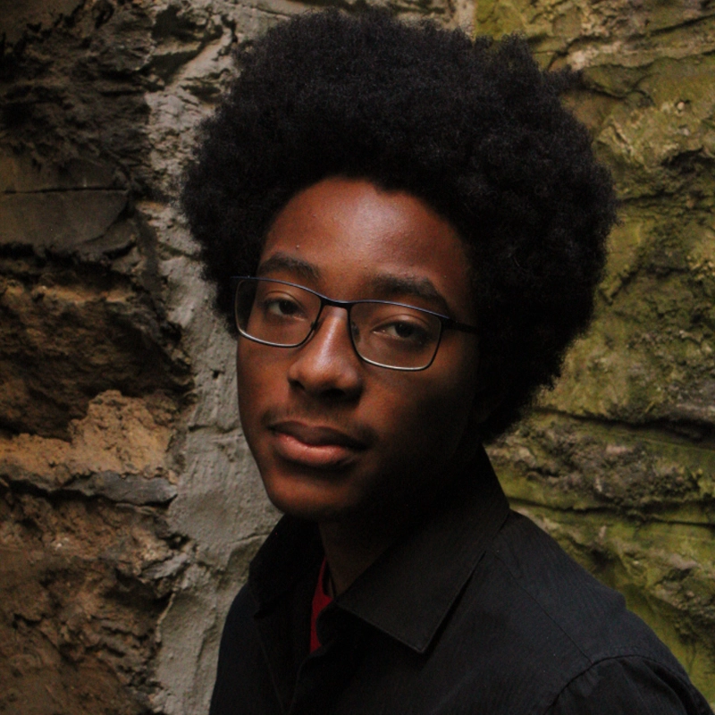 A young person with an afro hairstyle and glasses, wearing a black shirt, stands before a rough, mossy stone wall. Ellis Foreman looks calmly at the camera with a neutral expression as soft, natural light falls over them.