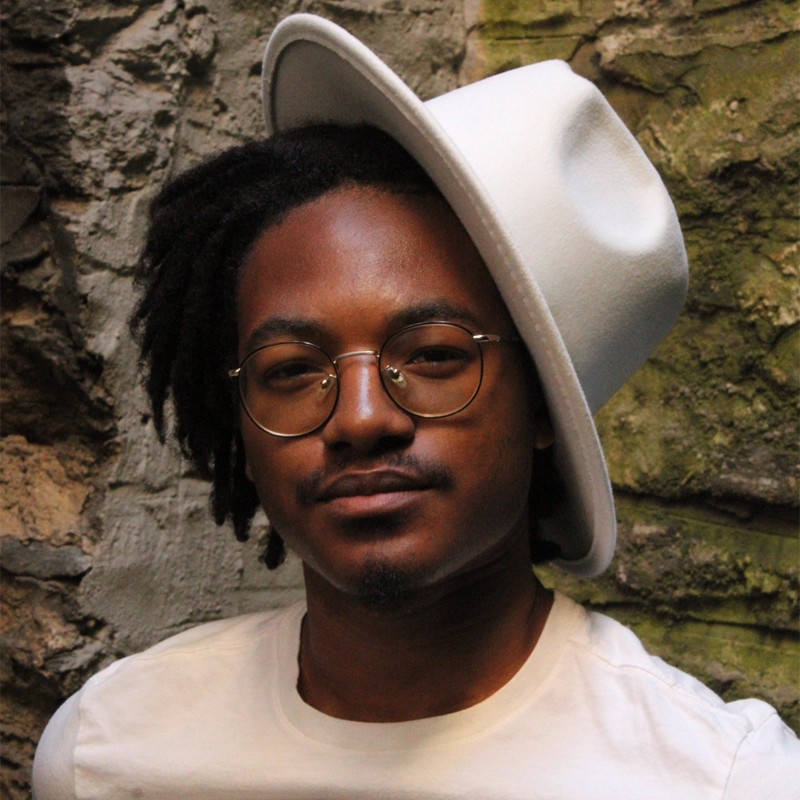 A person with short dreadlocks and a mustache, resembling Jaden Foreman, wears round glasses and a white hat tilted to the side. They are dressed in a white shirt, standing against a textured stone wall with green moss, warmly lit to highlight their face.