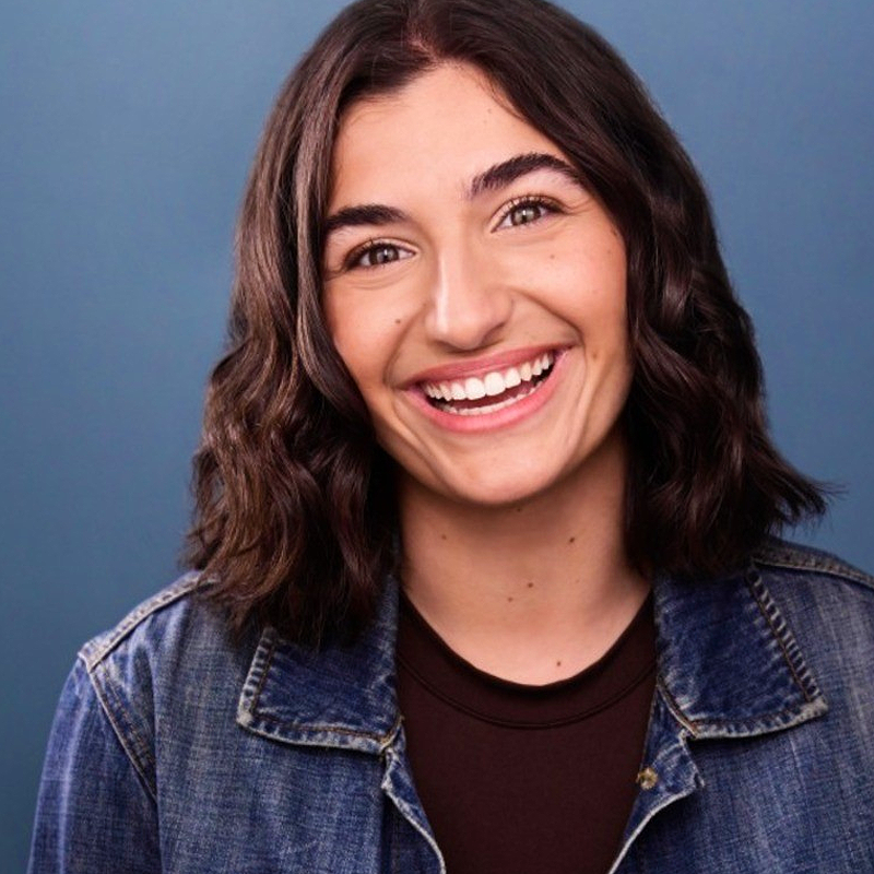 A young woman with wavy dark brown hair smiles widely. She wears a blue denim jacket over a dark top, and stands against a solid blue background. Her expression is cheerful and friendly, and she looks directly at the camera.