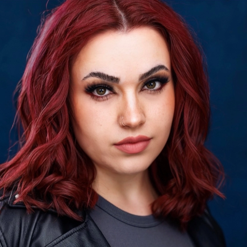 Grace Whetstone, a young woman with wavy, shoulder-length dark red hair, poses confidently against a dark blue background. She flaunts bold eyebrows, defined eye makeup, nude lipstick, a nose ring, and a black leather jacket.