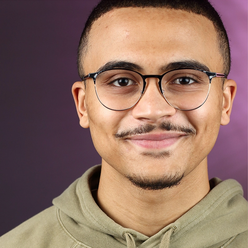 Gabe Woodard, a young man with short hair and glasses, smiles slightly at the camera. He wears a light olive hoodie against a dark purple gradient background, softly lit by pink on one side.