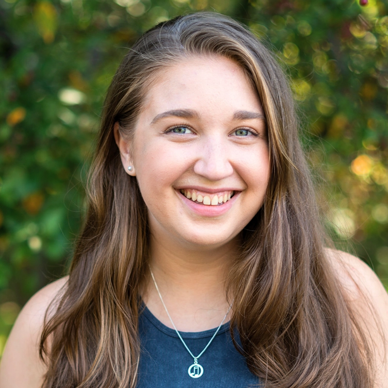 A young woman with long brown hair and blue eyes smiles at the camera. She is wearing a sleeveless dark top and a silver necklace with a round pendant. The lush, softly blurred greenery hints at photographer Joey Rodriguez’s signature style.