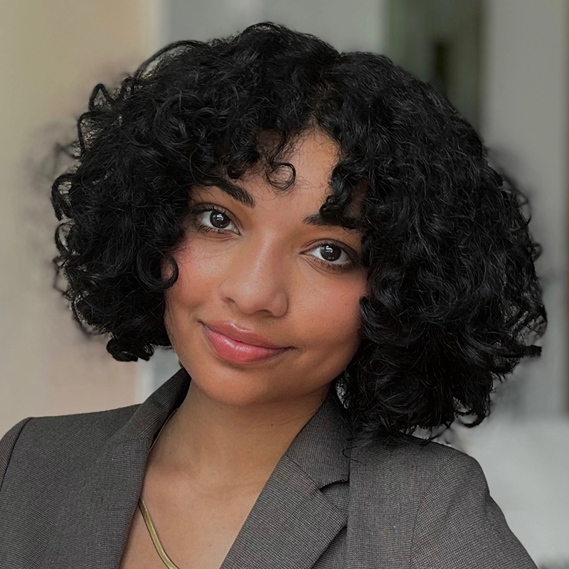 A person with short, curly black hair and medium skin tone smiles softly. Elsa Rose McIntyre Córdoba wears a gray blazer, a simple gold necklace, and natural makeup. The softly blurred background highlights her face.