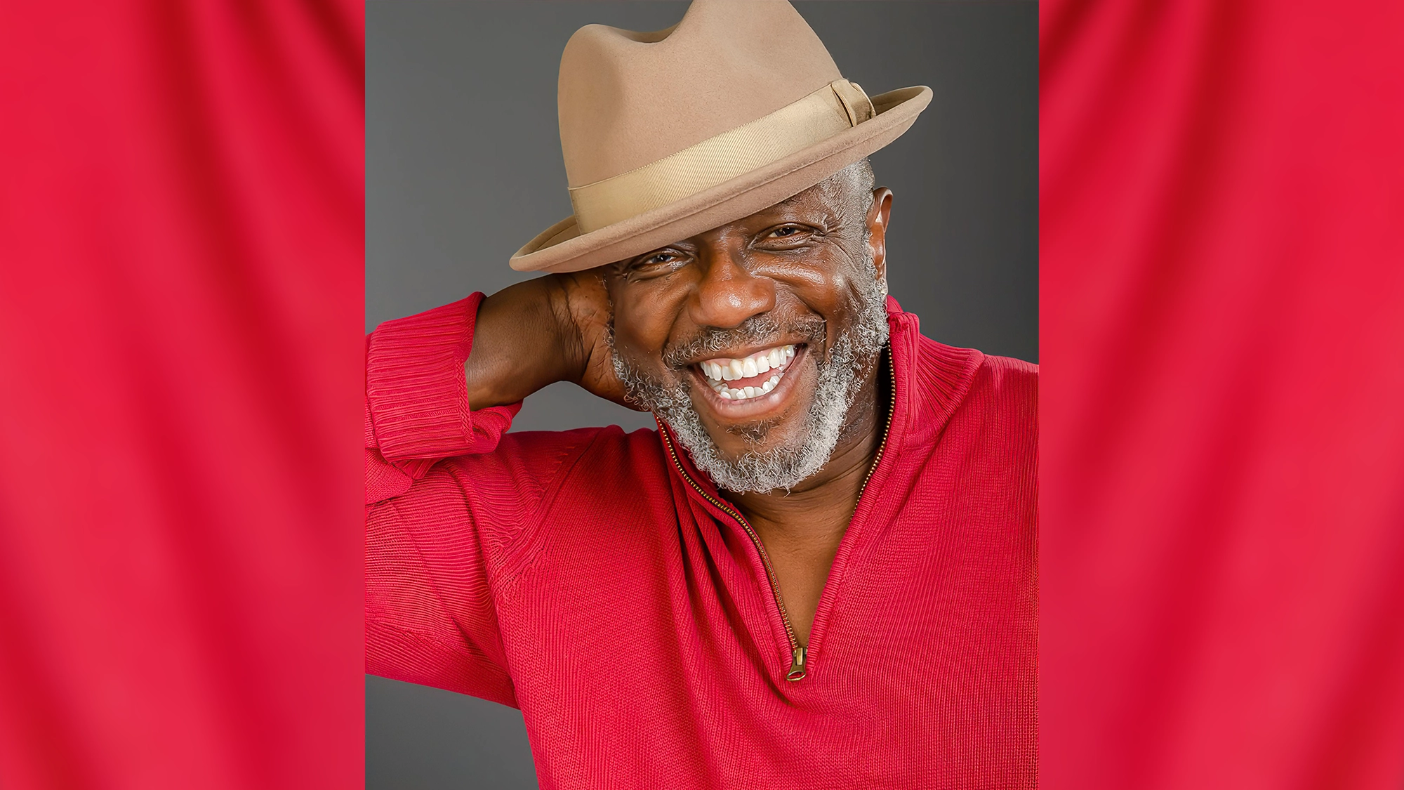 A joyful older man, George Young, with a gray beard smiles broadly, showing his teeth. He wears a tan fedora tilted to the side and a red zip-up sweater, posing with one hand behind his head against a gray background and red curtains.