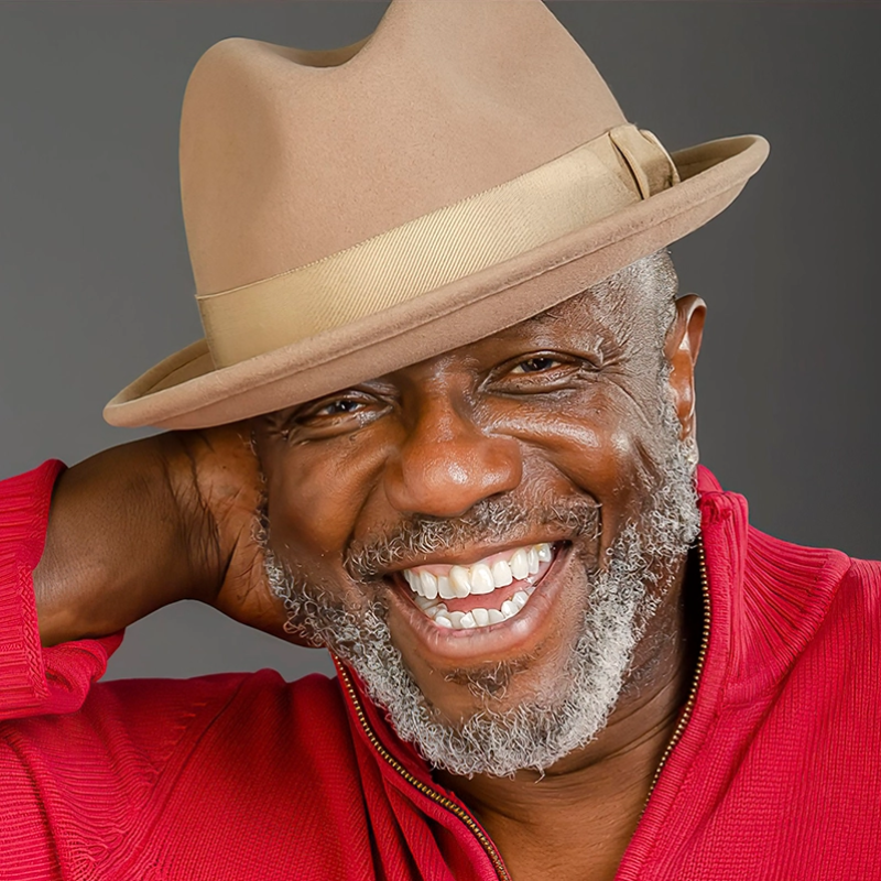 A smiling older man with a grey beard wears a tan fedora with a beige band and a red sweater. He tilts his hat back with one hand behind his head, showing bright white teeth. The background is plain gray.