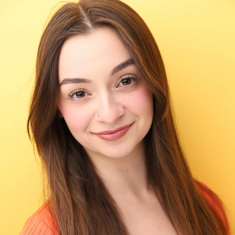 Young woman with straight brown hair and a nose stud smiles softly at the camera. She wears a peach top and light makeup, with defined brows and pink blush. The background is a solid, vibrant yellow, creating a warm, cheerful atmosphere.