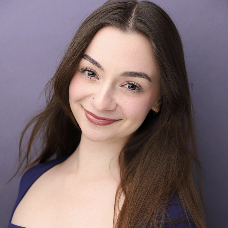 A young woman with long brown hair and fair skin smiles softly at the camera. She has dark eyebrows, brown eyes, and is wearing a dark blue top. The background is a smooth, lavender-purple color. Her head is slightly tilted to the left.