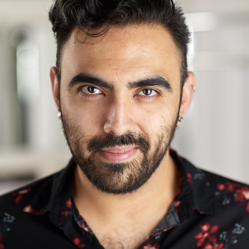 Alan González, a man with dark hair, a full beard, and mustache, stares confidently at the camera. He wears a black shirt with a red floral pattern and small hoop earrings. The softly blurred background draws focus to his expressive face and direct gaze.