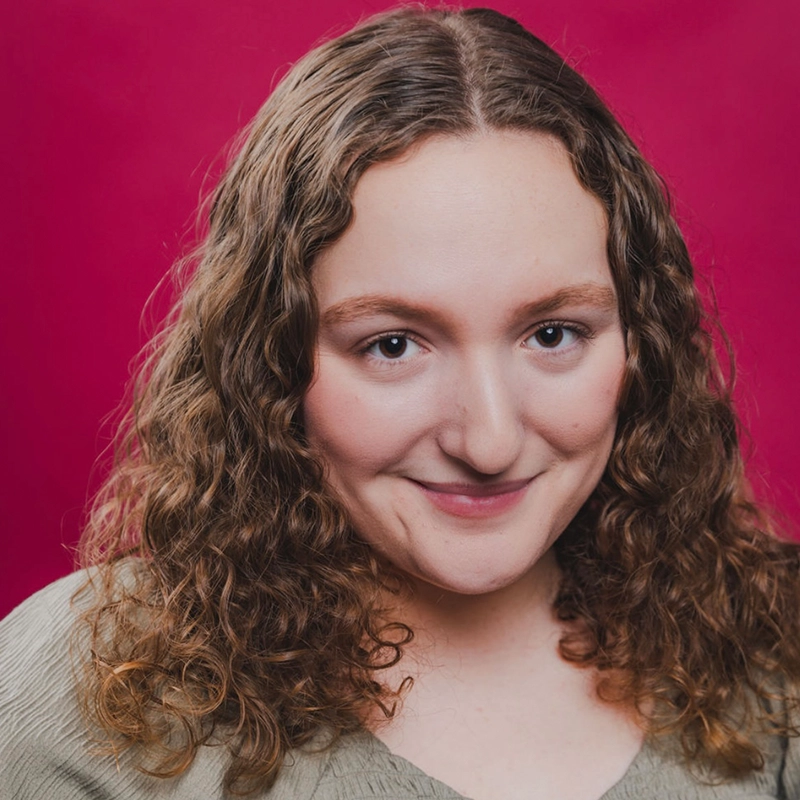 Grace Whetstone, with long, brown, curly hair and fair skin, smiles gently at the camera in a light green top against a solid magenta background.