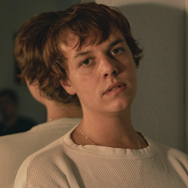 A person with short, tousled brown hair and light skin gazes softly at the camera. Jordan Graham wears a white textured shirt and a thin necklace. Their reflection appears in a mirror behind them, with the softly lit background out of focus.