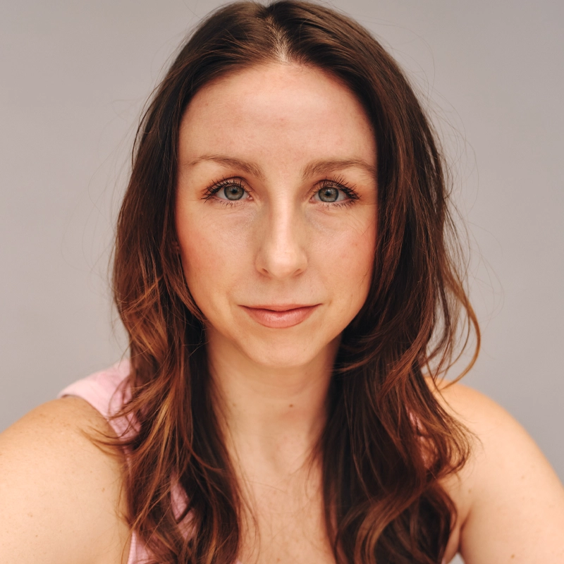 A woman with long, wavy brown hair and light skin looks directly at the camera with a neutral expression. She has blue-green eyes, subtle makeup, and wears a light pink top. The background is plain and light-colored.