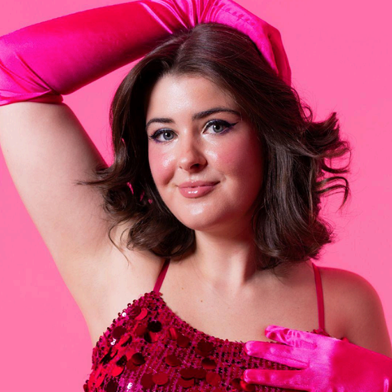 Gillian Gurganus, a woman with short, wavy brown hair, poses against a bright pink background. She wears long, shiny pink gloves and a sparkling red sequin top, smiling softly with her right arm raised above her head.