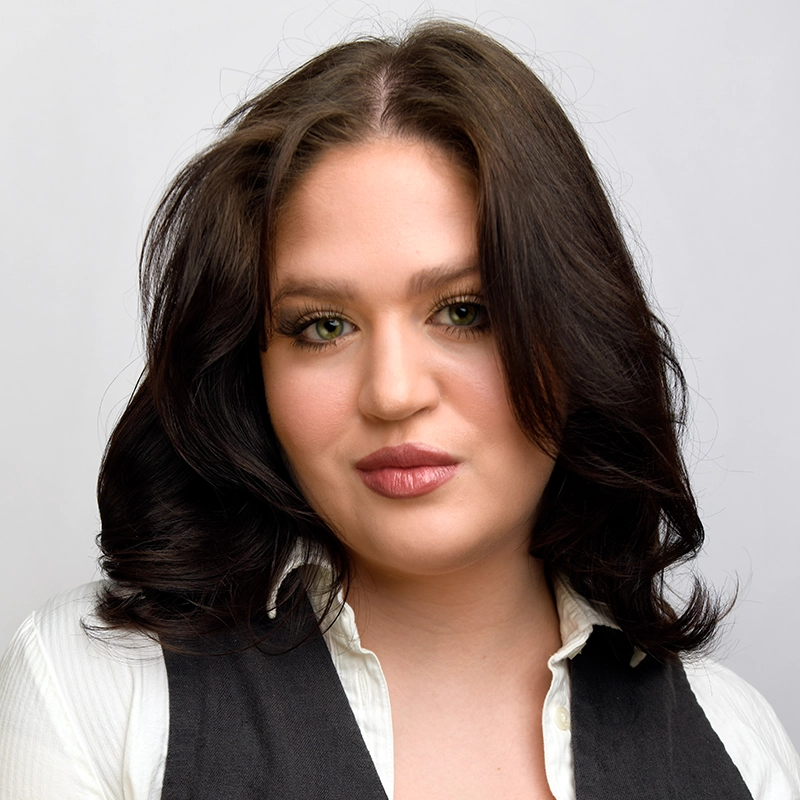 A young woman with fair skin and shoulder-length dark brown hair, styled in loose waves, poses against a plain, light background. She wears a white collared shirt with a dark vest, echoing Kitana's elegance, and has neutral makeup, full lips, and green eyes.