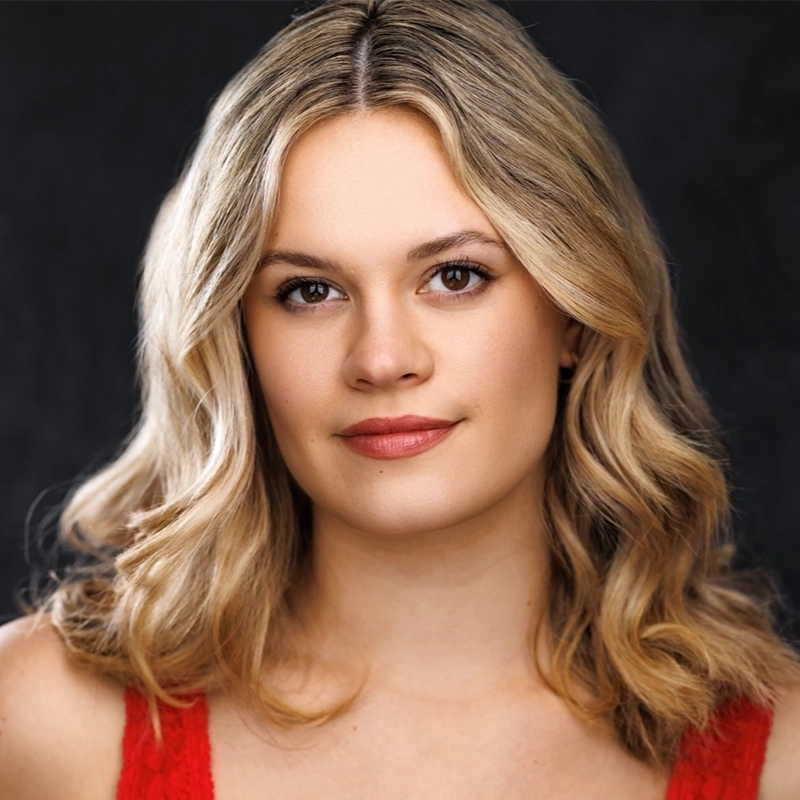 Grace Whetstone, a young woman with wavy, shoulder-length blonde hair and fair skin, looks directly at the camera with a slight smile. She wears a sleeveless red top and natural makeup, set against a dark, blurred background.
