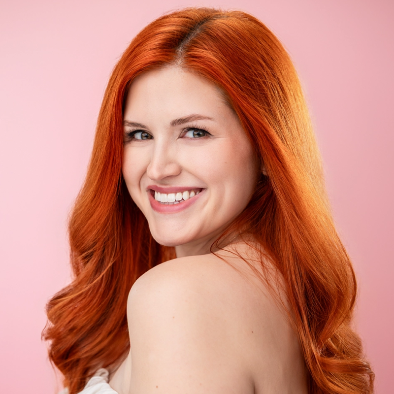 A woman with long, wavy red hair smiles over her shoulder at the camera. She has fair skin and is wearing a white, off-shoulder top. The background is a solid, soft pink color, and the lighting is bright and even.