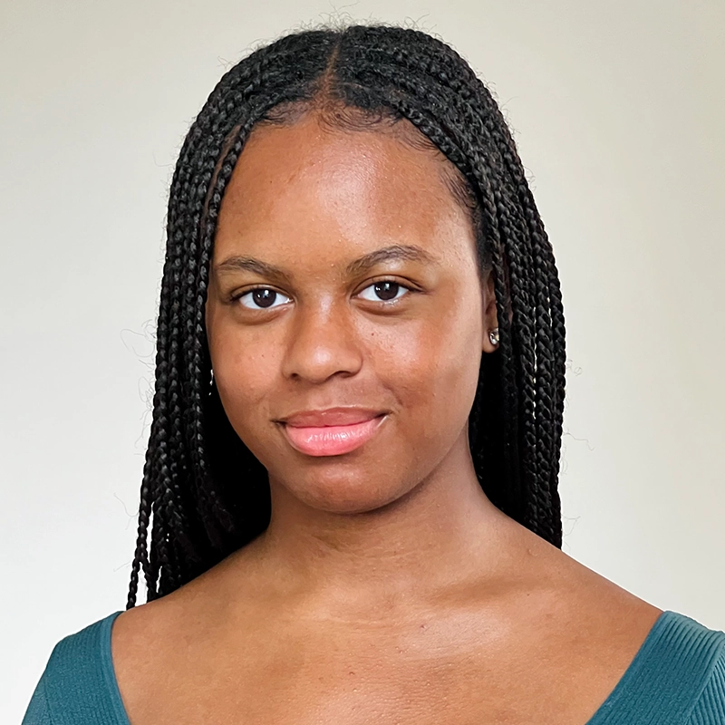 Anjalee Hardy, a young woman with long, neat box braids, smiles softly at the camera. She has smooth, medium-brown skin and wears a teal top against a plain, light background that keeps the focus on her face.
