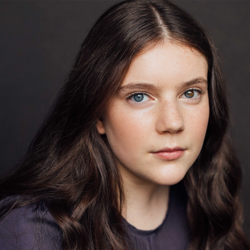 A young girl with long, wavy brown hair and light skin gazes directly at the camera. She has blue eyes and a calm, neutral expression. She wears a dark top, and the background is plain and dark, highlighting her features.