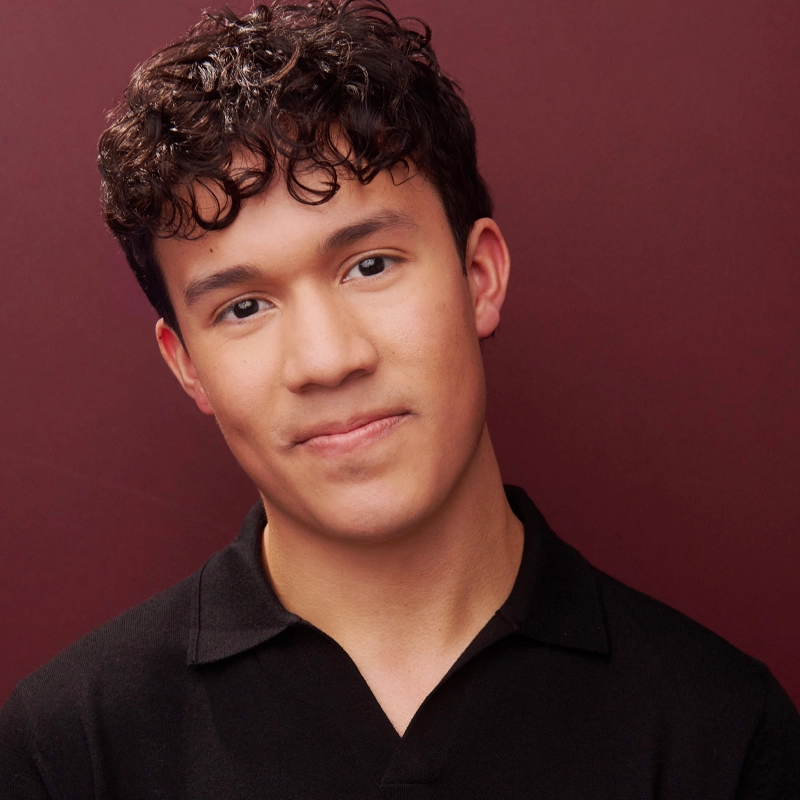 A young person with short, curly dark hair and light skin smiles softly at the camera. Kaden Hawkins wears a black collared shirt and stands before a plain maroon background. The image is well-lit, radiating a warm and friendly tone.