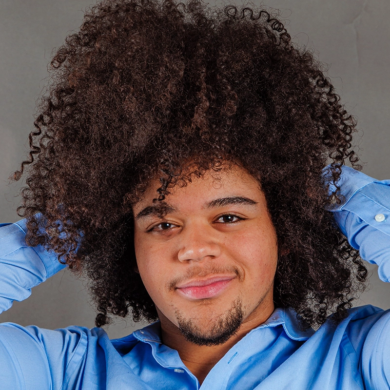 A young man with curly, voluminous dark hair smiles gently while holding his hair up with both hands. Styled by Coral Rosario-Laboy, he wears a light blue button-up shirt and appears confident and relaxed against a neutral gray background.