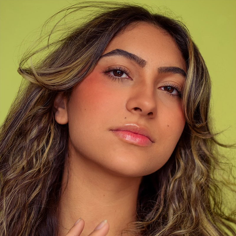 A young woman with wavy, highlighted brown hair stands against a light green background. Channeling the elegance of Isabella Henao, she has strong eyebrows, subtle eyeshadow, rosy blush, and glossy lips, gazing calmly at the camera with a relaxed expression.