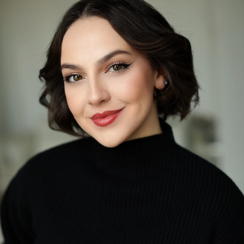 Reese Henrick, a woman with short dark hair and brown eyes, smiles softly. She wears winged eyeliner, red lipstick, and a black ribbed turtleneck sweater. The softly blurred background highlights her face as the main focus of the image.