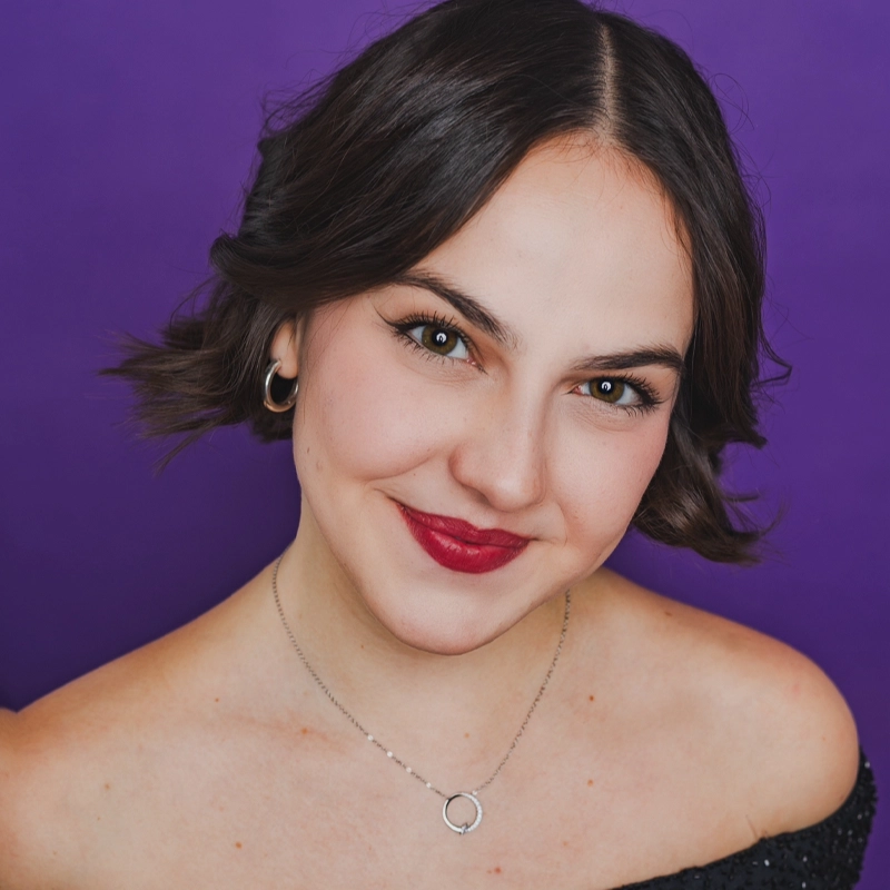 Reese Henrick smiles warmly against a solid purple background. She has short, dark wavy hair, silver hoop earrings, a delicate necklace with a circular pendant, and a black off-the-shoulder top. Her bold red lipstick and defined brows stand out.