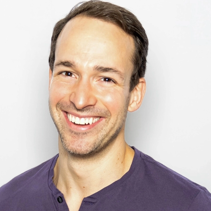 Christopher Herr, a smiling man with short brown hair, wears a purple shirt with buttons at the collar. Facing the camera against a plain, light background, he is brightly lit, highlighting his cheerful expression and even skin tone.