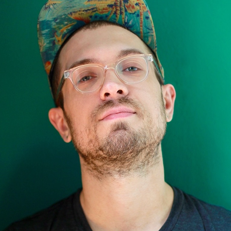 A person with light skin, clear glasses, and facial stubble stands against a green background. They wear a colorful, patterned cap and a dark t-shirt. Their head is slightly tilted, and they have a neutral, relaxed expression.