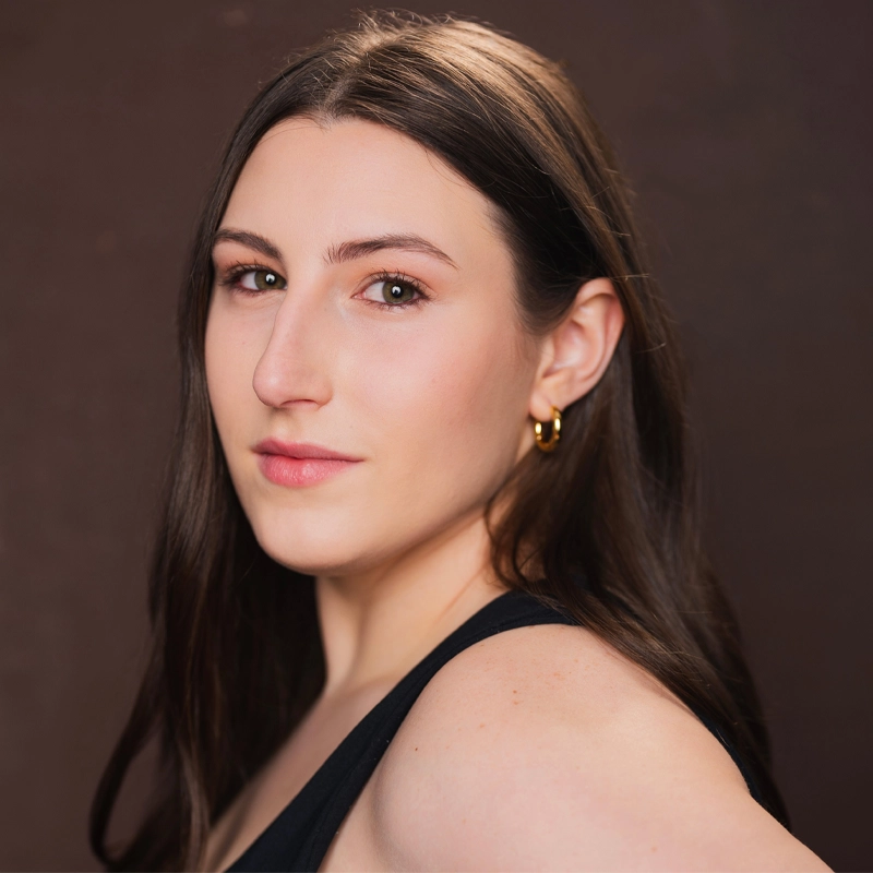 A young woman with long straight brown hair and light skin looks over her shoulder at the camera. She wears a black sleeveless top and gold hoop earrings. The background is plain and dark brown, highlighting her natural makeup and calm expression.