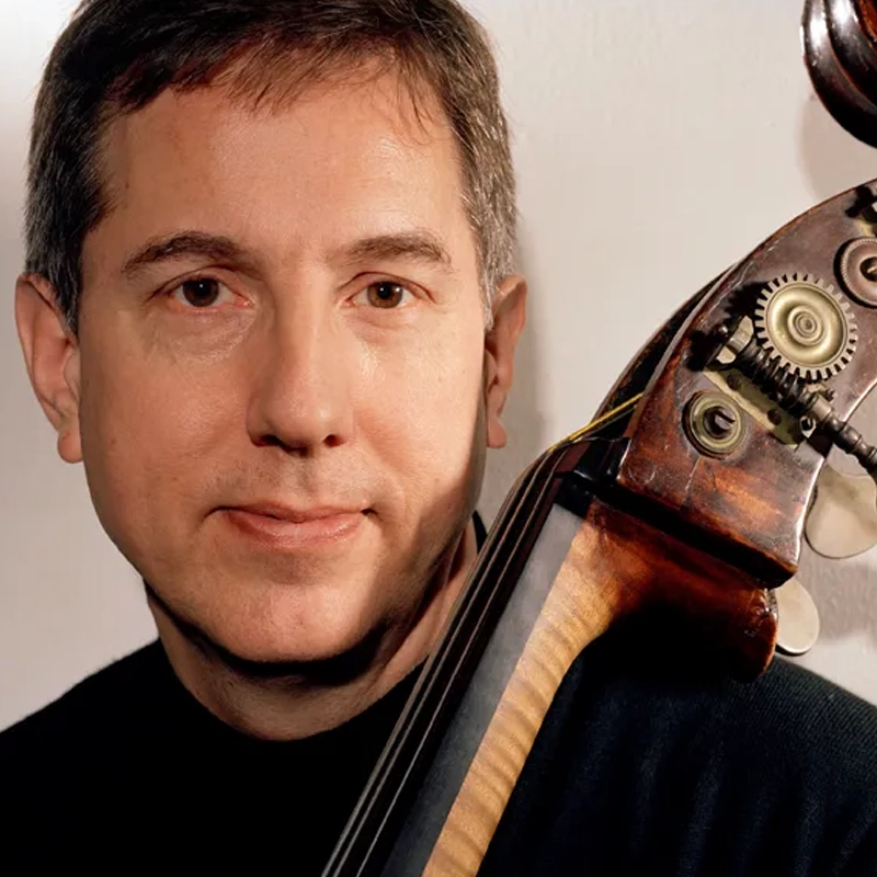 A man with short brown hair, wearing a black shirt, poses with a double bass. The scroll and tuning pegs of the instrument are visible beside his face. The background is plain and light-colored.