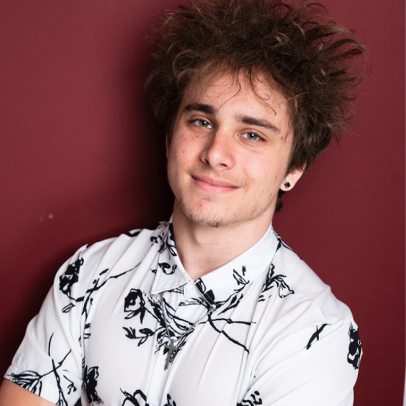 A young person with messy brown hair and light skin smiles slightly, arms crossed. They wear a white shirt with black abstract patterns and a small black earring in one ear. The solid maroon background highlights their Ian McCaslin-inspired style.