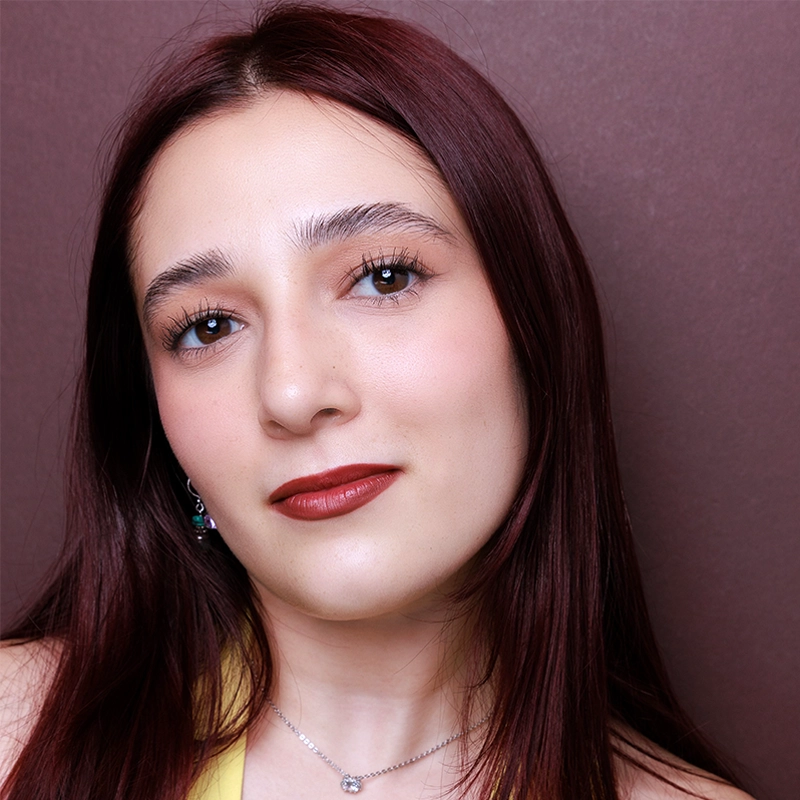 A young woman, Ingrid Orozco, with straight, reddish-brown hair poses in front of a maroon background. She has fair skin, brown eyes, and red lipstick. She wears a yellow top, silver necklace, and a small nose piercing.