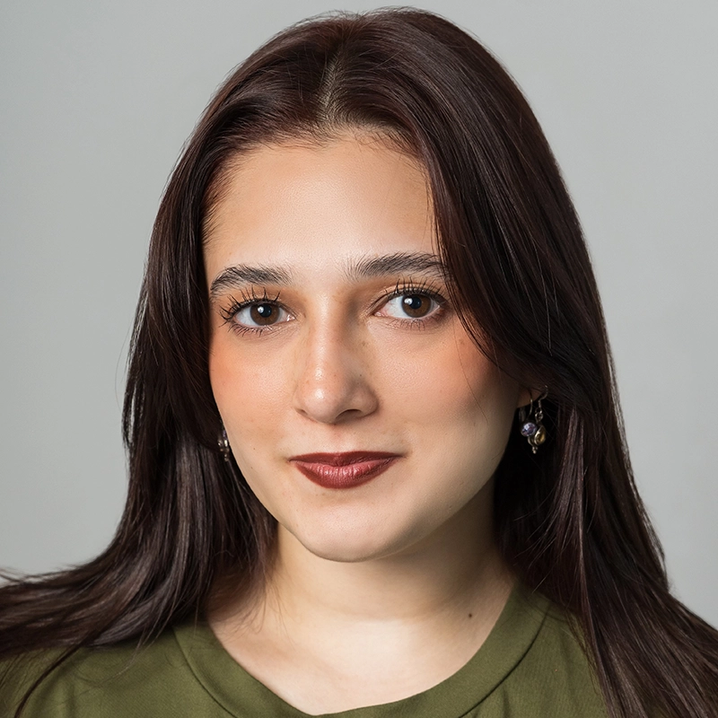 A young woman with long, dark brown hair poses against a light gray background. She has fair skin, brown eyes, and wears subtle makeup with brown lipstick. Wearing a green top and small earrings, she gazes calmly at the camera, embodying Raíces.