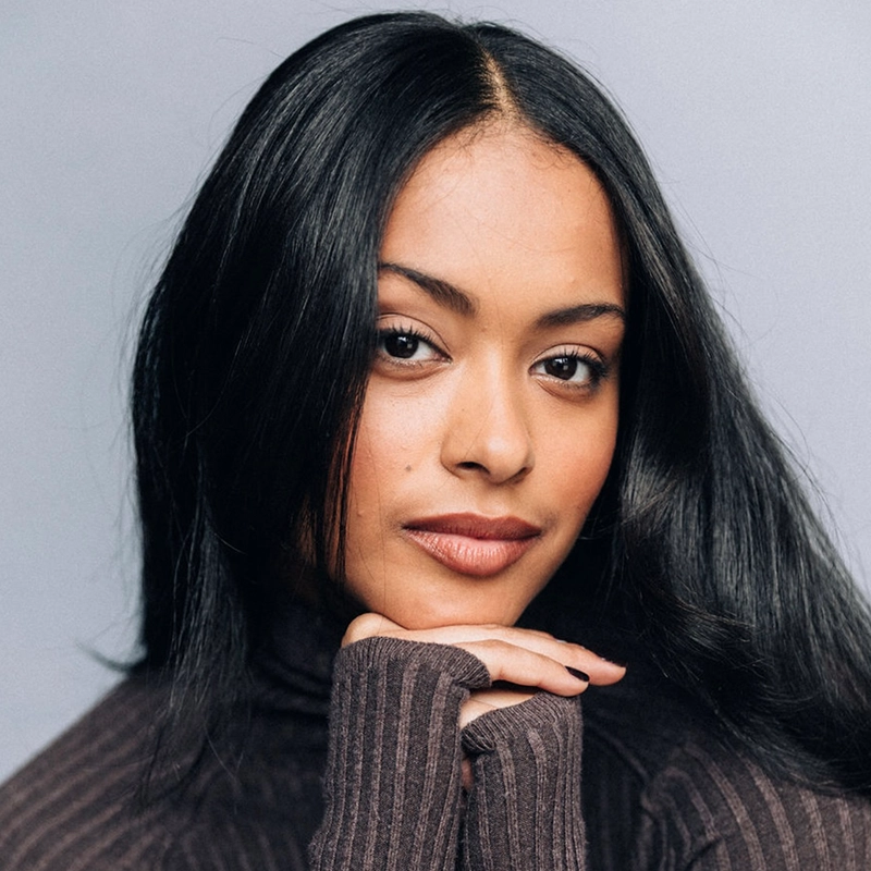 Isabel Gonzalez, with long, straight black hair and smooth skin, poses against a plain gray background. She wears a ribbed, dark brown turtleneck sweater, resting her chin on her hand with a subtle, confident smile and calm, direct gaze.