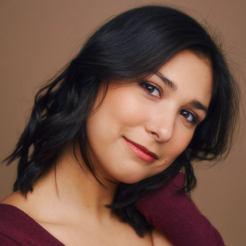 Grace Whetstone, with shoulder-length dark hair, tilts her head and smiles gently at the camera. She wears a burgundy top and natural makeup with rosy lips, set against a soft beige background for a warm, inviting feel.