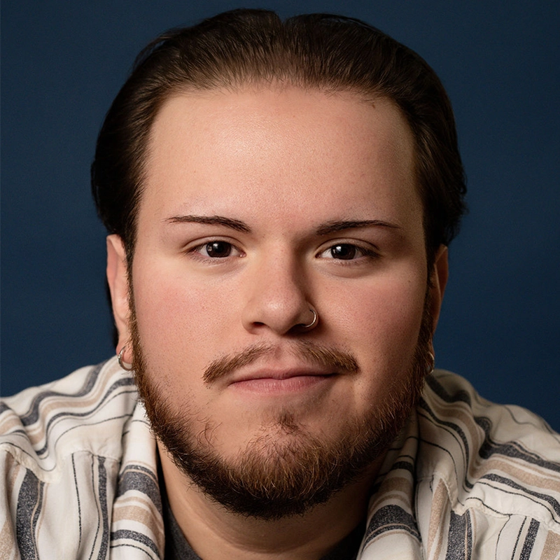 A young person with short dark hair, a trimmed beard, and a nose ring looks directly at the camera, smiling slightly. They wear a striped shirt over a dark top. The background is a solid dark blue.