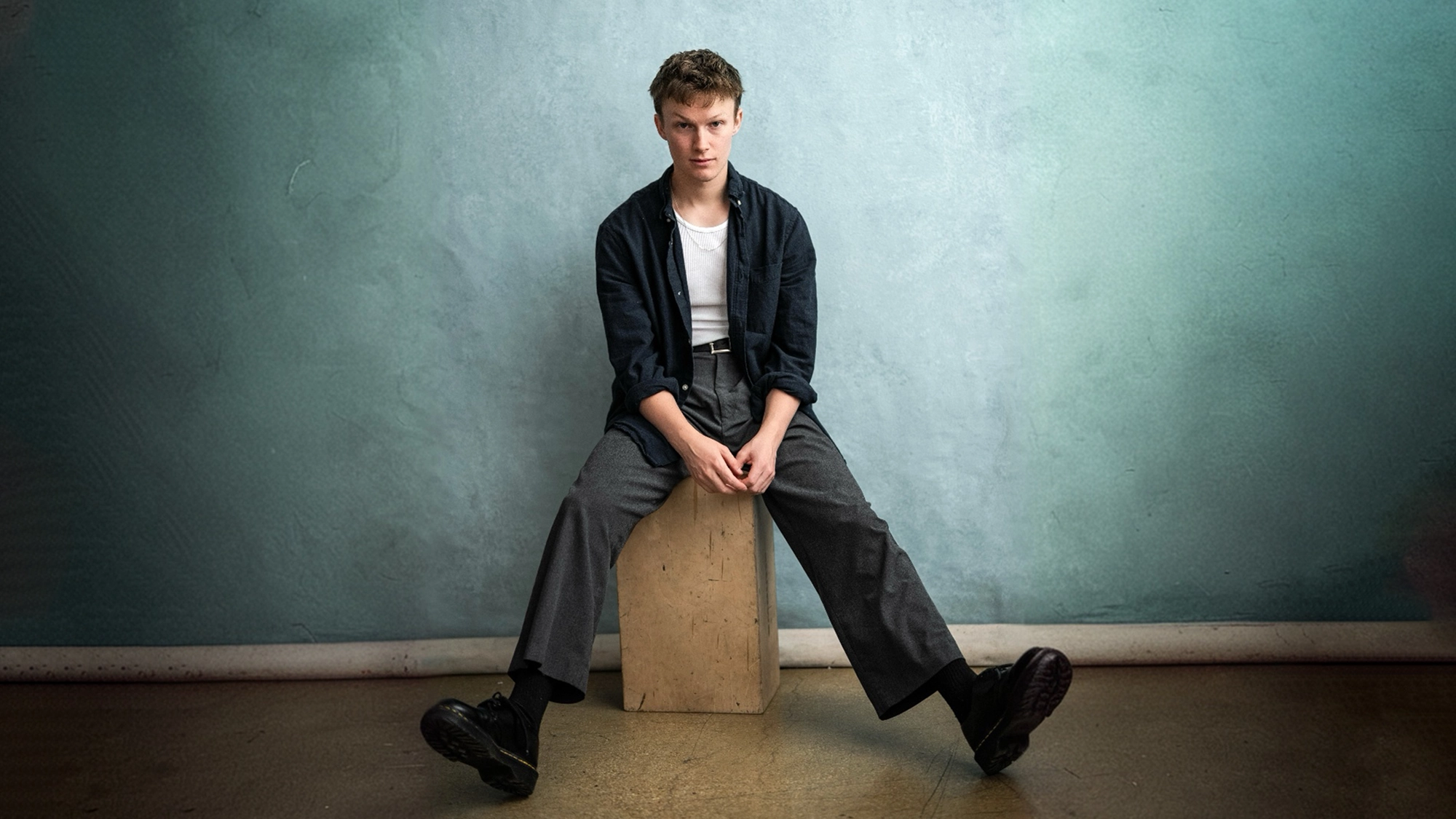 Jack Roden site image 26 A young man with short hair sits on a wooden cube against a textured teal wall. He wears a dark shirt over a white T-shirt, gray wide-leg pants, and black shoes. Jack Roden rests his arms on his knees and looks directly at the camera with a neutral expression.