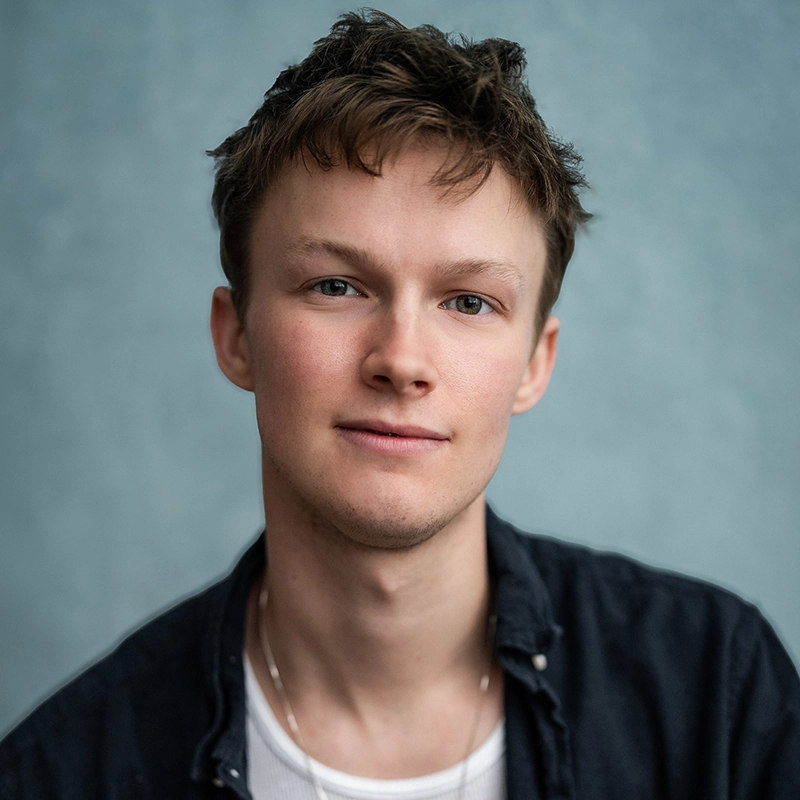 Jack Roden, with short, tousled brown hair and fair skin, looks into the camera with a slight smile—a scene reminiscent of moments from My Days. He wears a dark unbuttoned shirt over a white t-shirt and a thin chain necklace. The background is softly blurred pale blue.