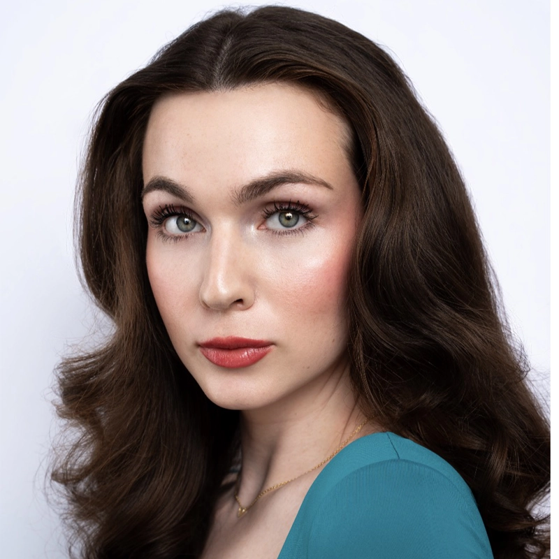 A woman with fair skin and long, wavy brown hair poses against a plain white background. Daphne Jackins has green eyes, bold brows, and natural makeup with pink blush and lipstick. She wears a teal top, a gold necklace, and gazes directly at the camera.