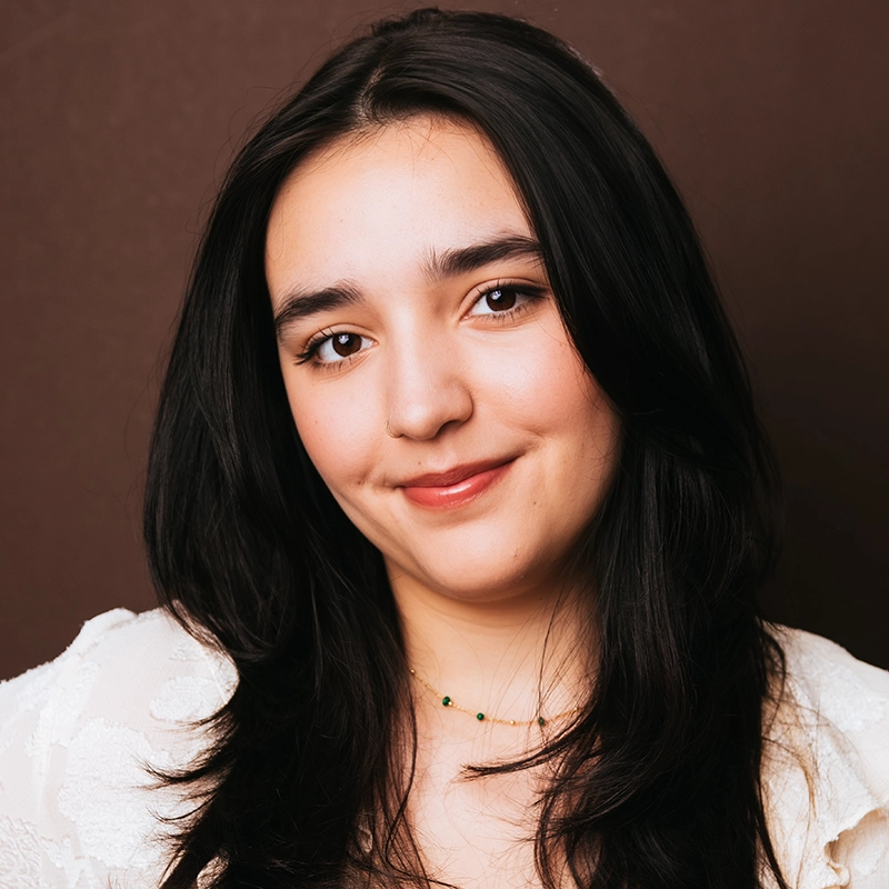 Grace Whetstone, a young woman with long black hair and fair skin, smiles gently at the camera. She wears a white textured top and a delicate beaded necklace. The background is plain brown, with warm, soft lighting enhancing her serene presence.