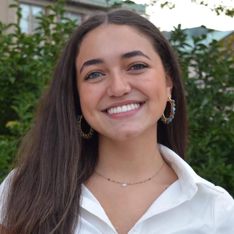 Jaden Natividad, a young woman with long brown hair, wearing a white collared shirt, hoop earrings, and a delicate necklace, smiles broadly at the camera. Green foliage and a building are blurred in the background.