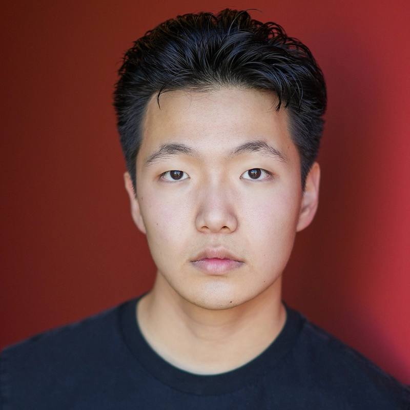 A young person with short, dark hair and fair skin looks directly at the camera with a neutral expression, wearing a black T-shirt. The background is a smooth gradient of deep red.